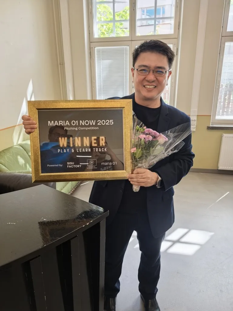 Same PSI representative smiling indoors, holding the framed "Maria 01 NOW 2025 Winner, Play & Learn Track" certificate and a bouquet of flowers