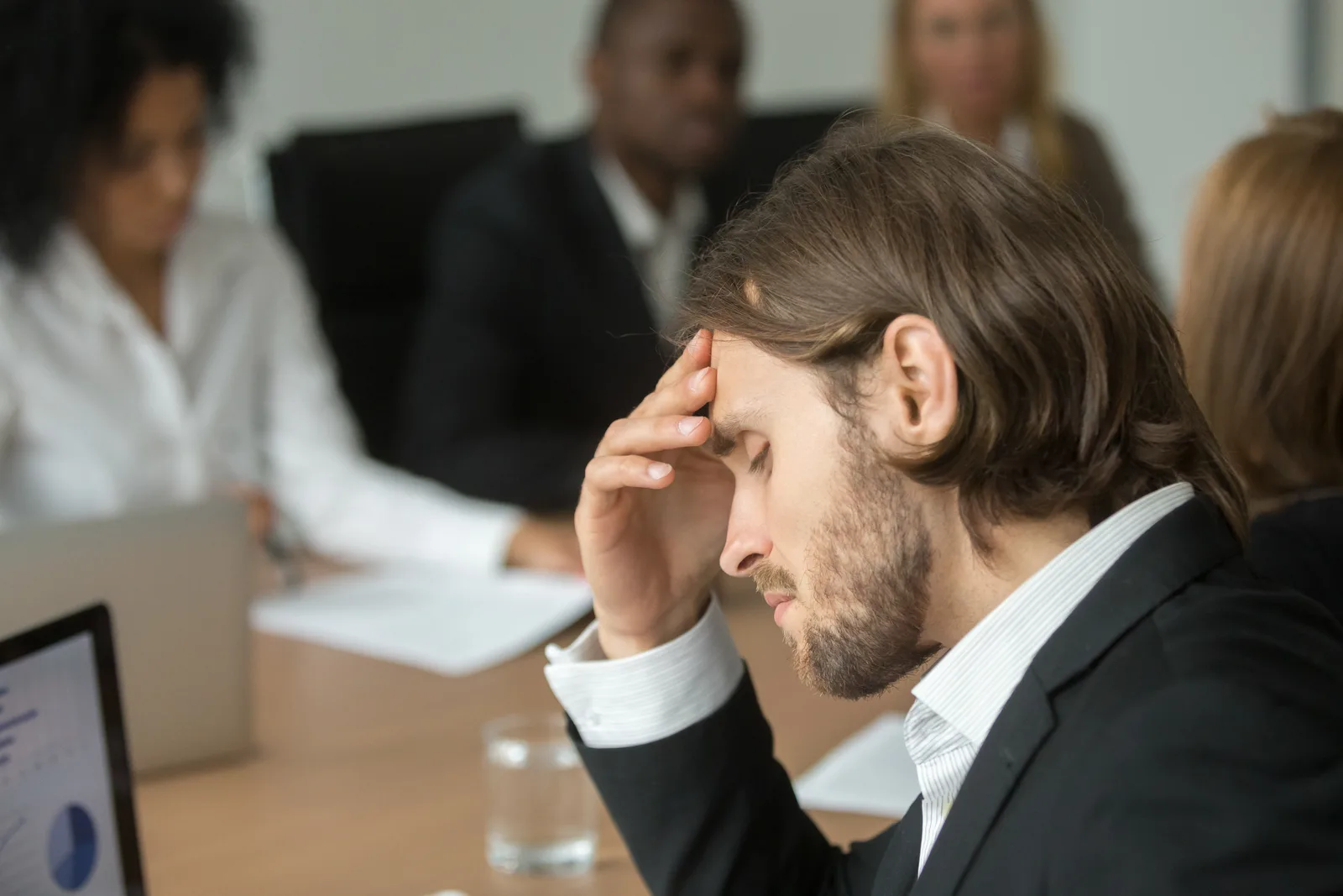 A man at a boardroom table with his eyes closed and his fingers pressed to his forehead, while colleagues talk around him and a laptop shows charts in the foreground.