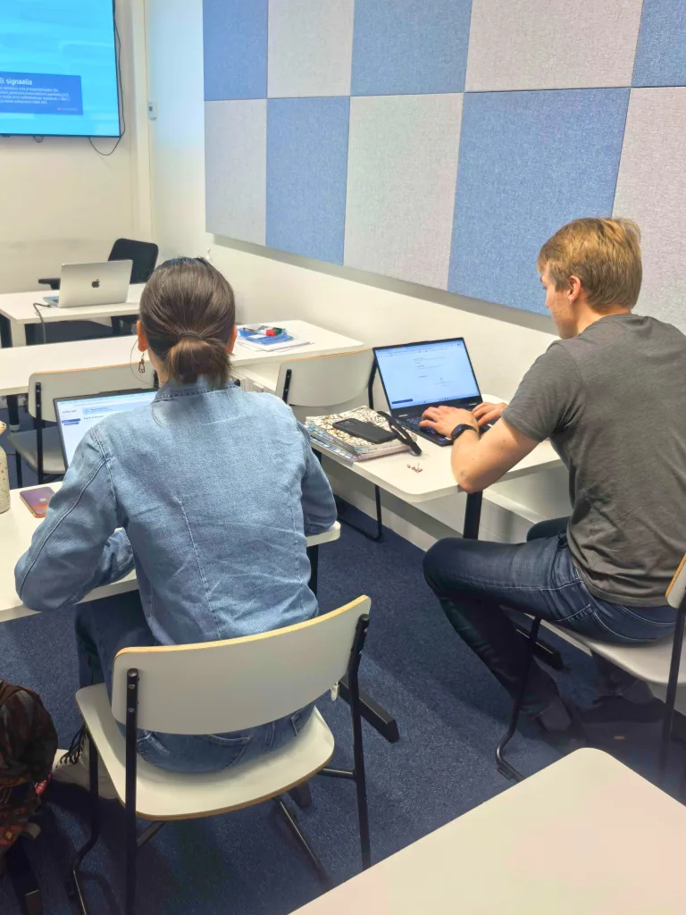 Photo of two people seated at desks working on laptops during a classroom demo session, with a presentation slide shown on a screen in the background
