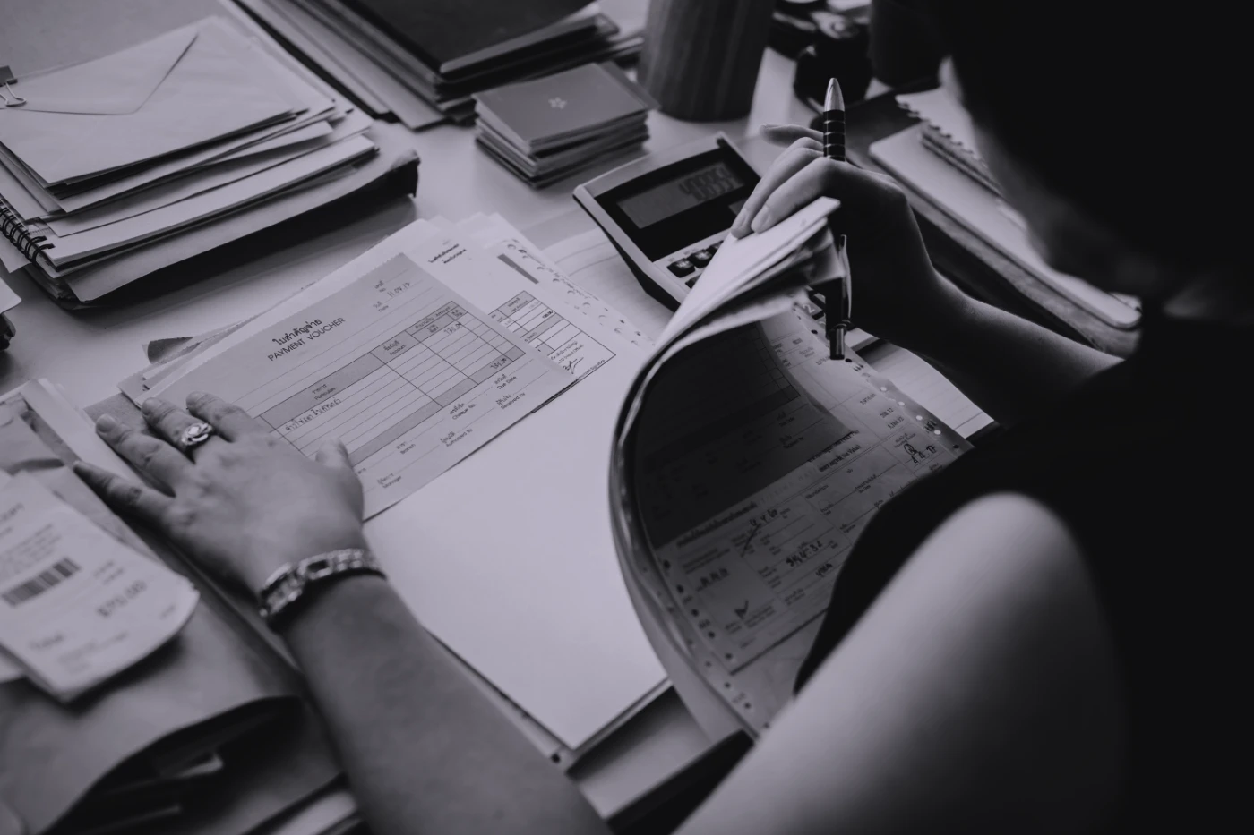 A clerk at a desk covered in paperwork: a payment voucher held flat under one hand, a carbon-copy ledger lifted with the other, a calculator, and stacks of filed documents to every side.